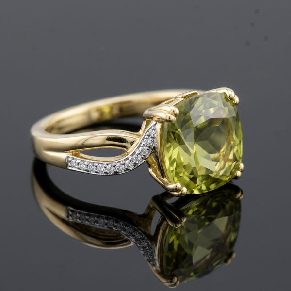 A yellow gold ring featuring a cushion-cut olive green gemstone, accented with small pavé-set clear stones on the band, shown on a reflective dark gray surface.