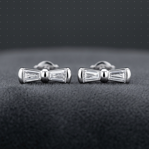 A pair of silver bow-shaped cufflinks with baguette-cut clear stones, displayed on a dark textured surface against a dark gray background.